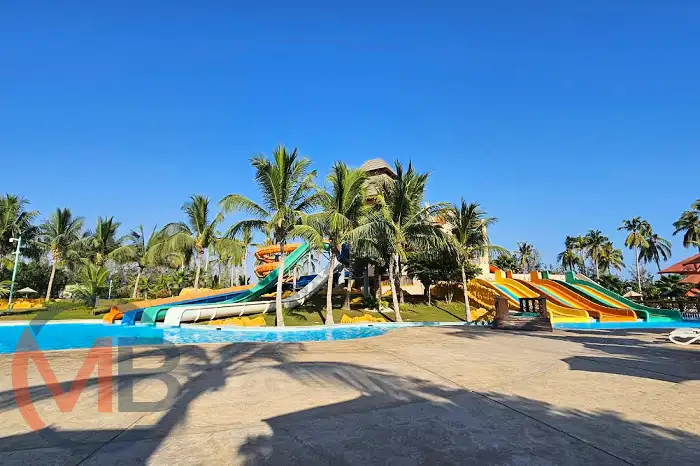 Visitors on colorful water slides and in a wave pool at Hawana Aqua Park, with palm trees and resort buildings in the Hawana Salalah resort area of Salalah (Wilayat), Dhofar Governorate